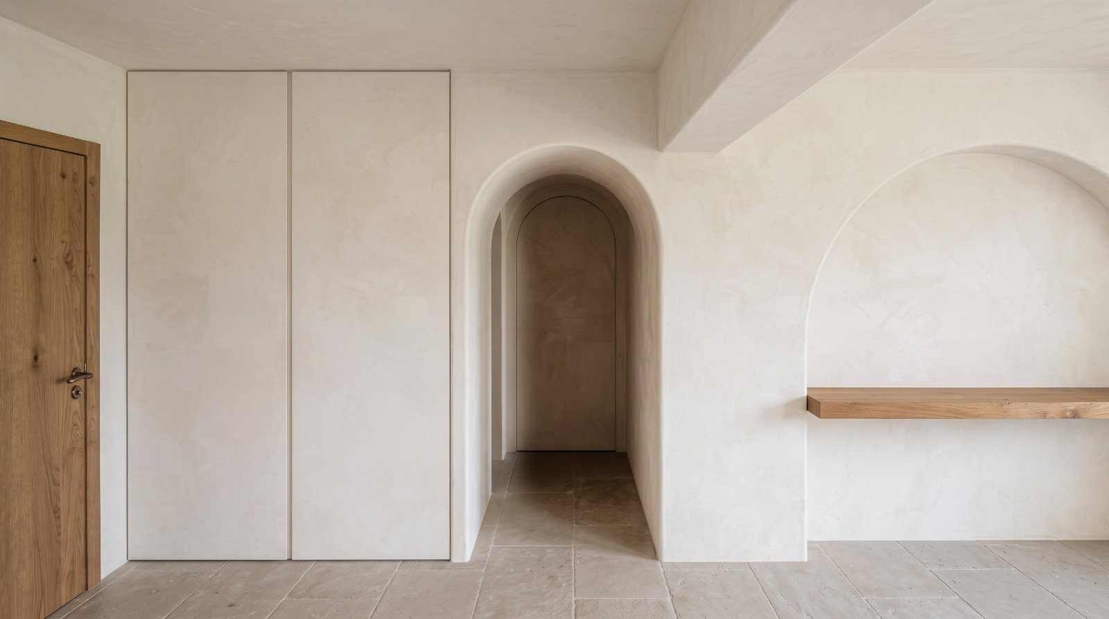 Lime-plastered room with flush cupboards, an arched passage and an arched niche with an oak shelf, on old stone flooring.