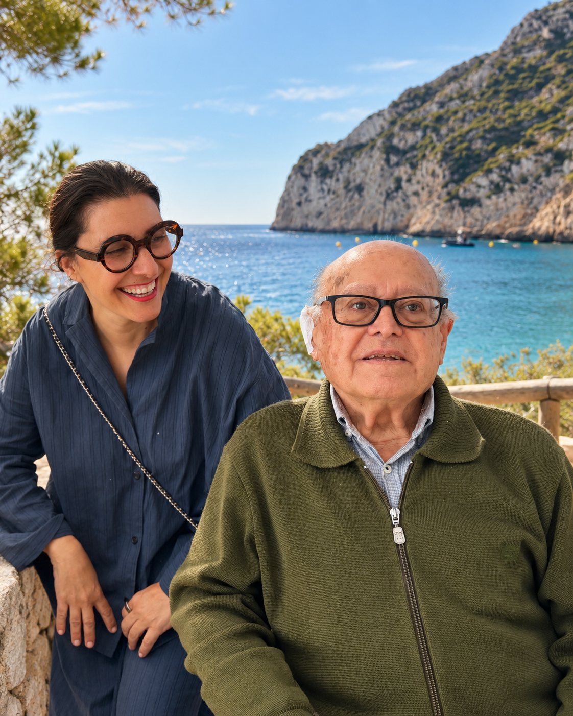 Don Salvador Ginestar Soler, seated, smiling. Beside him Jessica Bataille laughs. Behind them, the sea and the cliff of La Granadella.