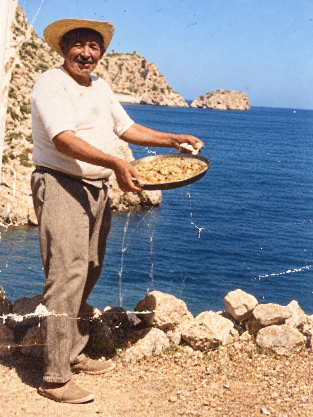 Old colour photograph: a man in a straw hat and espadrilles smiles, holding a paella by the sea, with an islet behind.